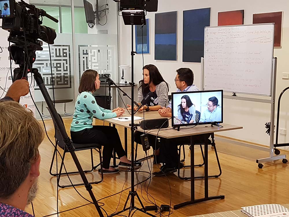 A group of four people seated around a table, engaged in a conversation at a public relations agency. A camera crew is filming them, with a monitor displaying the participants. The background shows a whiteboard with writing, large abstract paintings, and a glass wall.