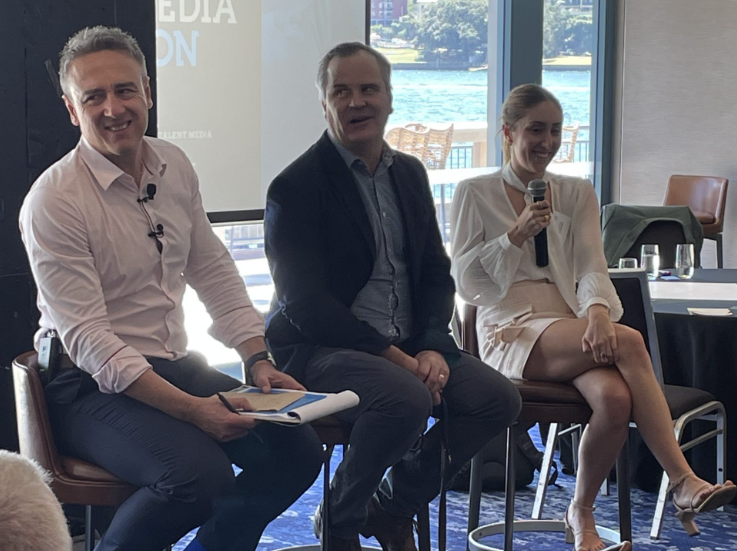 Three individuals are seated on stools in front of a room, engaged in a panel discussion. The man on the left holds a notebook, the man in the center is speaking, and the woman on the right from a public relations agency is holding a microphone and smiling. A large window in the background displays a waterfront view.