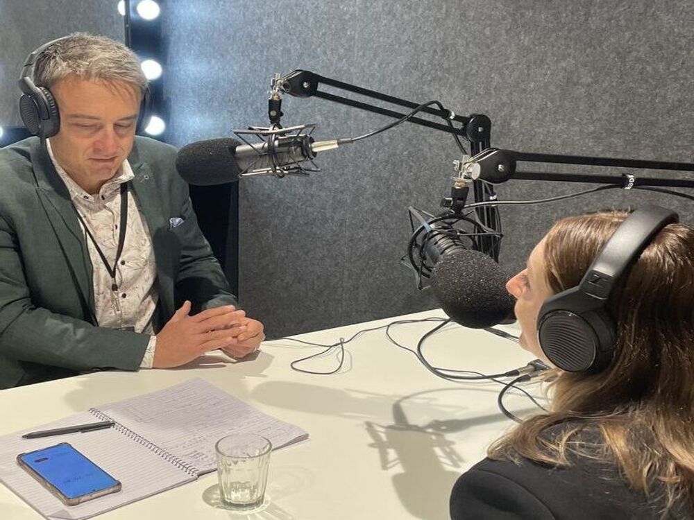 Two people wearing headphones and sitting across from each other in a podcast studio at a public relations agency. Each has a microphone in front of them on adjustable arms. There are notebooks and a smartphone on the table. The person on the left is speaking, while the person on the right listens attentively.