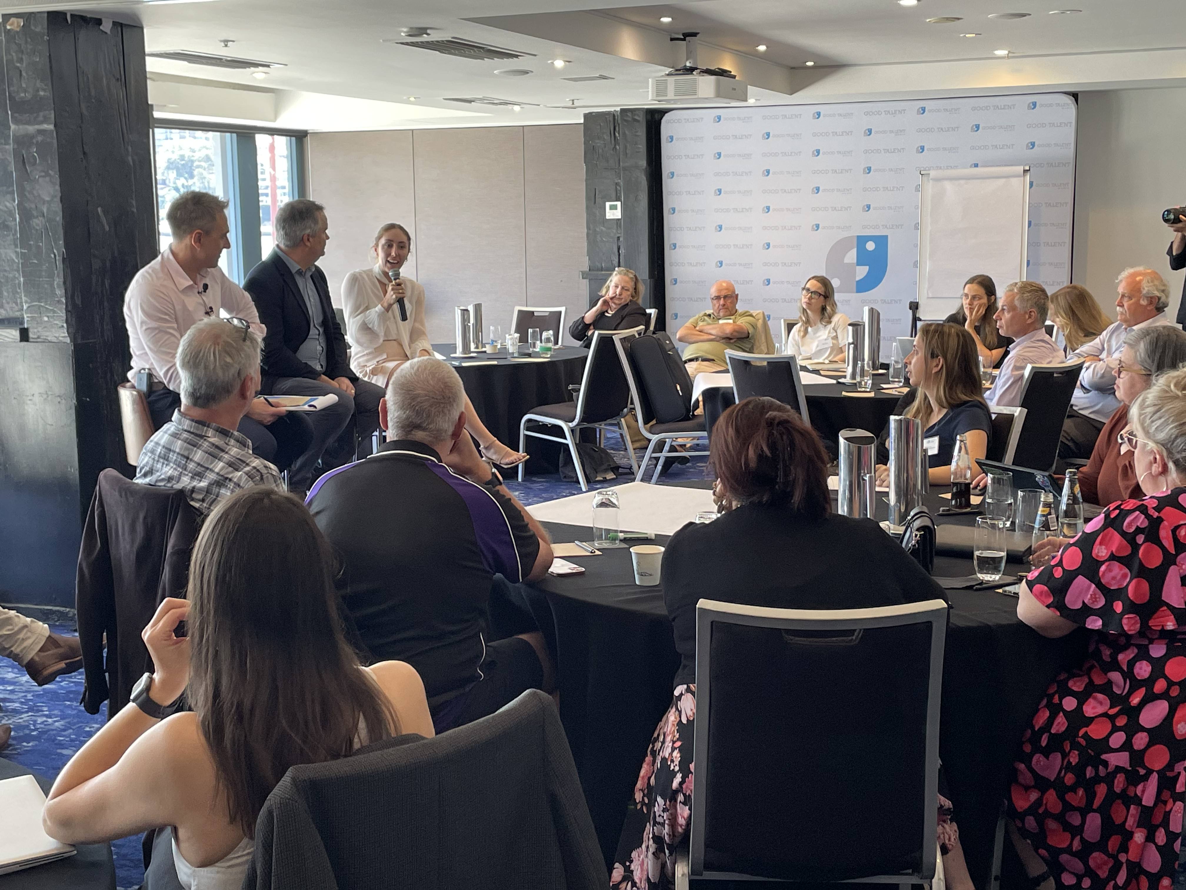 A group of people are gathered in a conference room, seated around tables. Three individuals from a public relations agency at the front are speaking, while the audience listens attentively. A backdrop with a logo featuring a comma can be seen, along with a whiteboard and a large window.