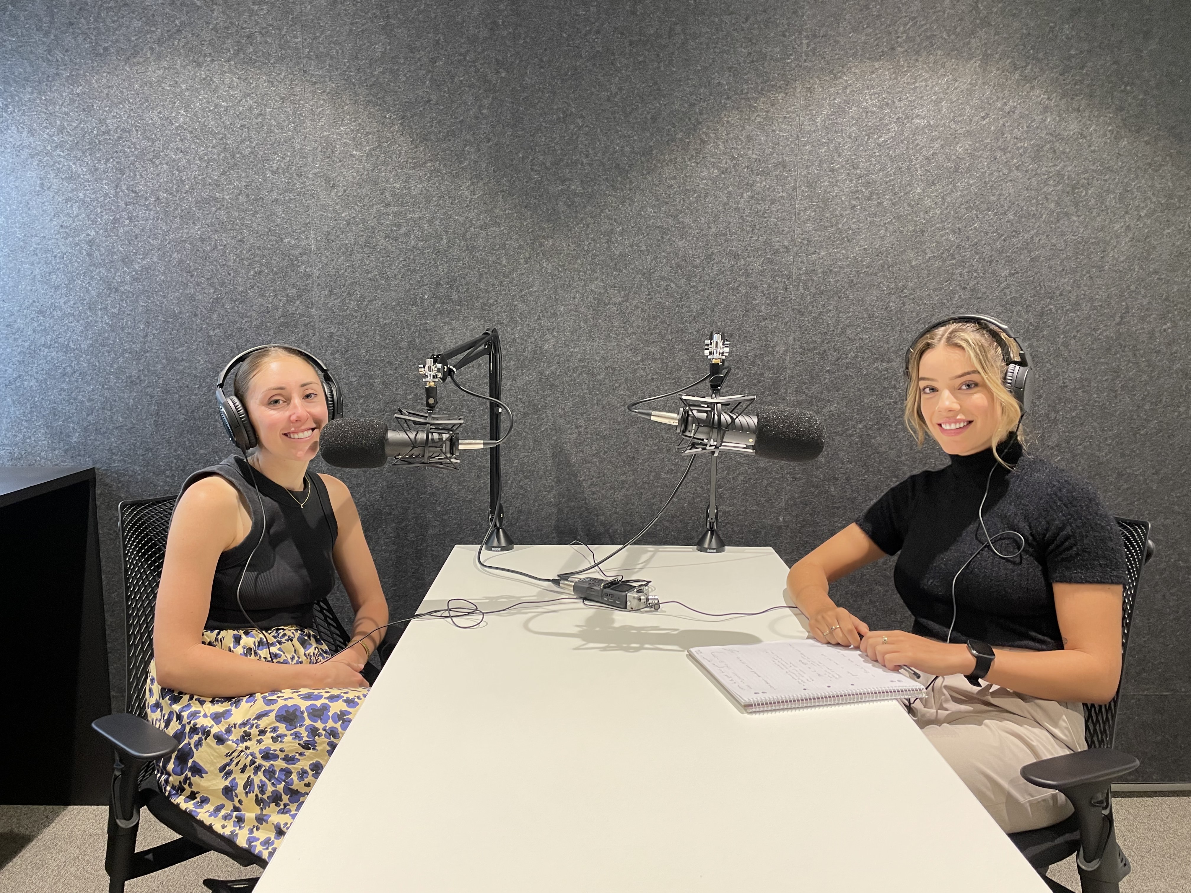 Two women sit in a podcast studio with professional microphones and headphones. Both are seated at a table, one holding a notebook. The background features soundproofing material, hinting at their affiliation with a public relations agency. They smile, ready for an engaging discussion.