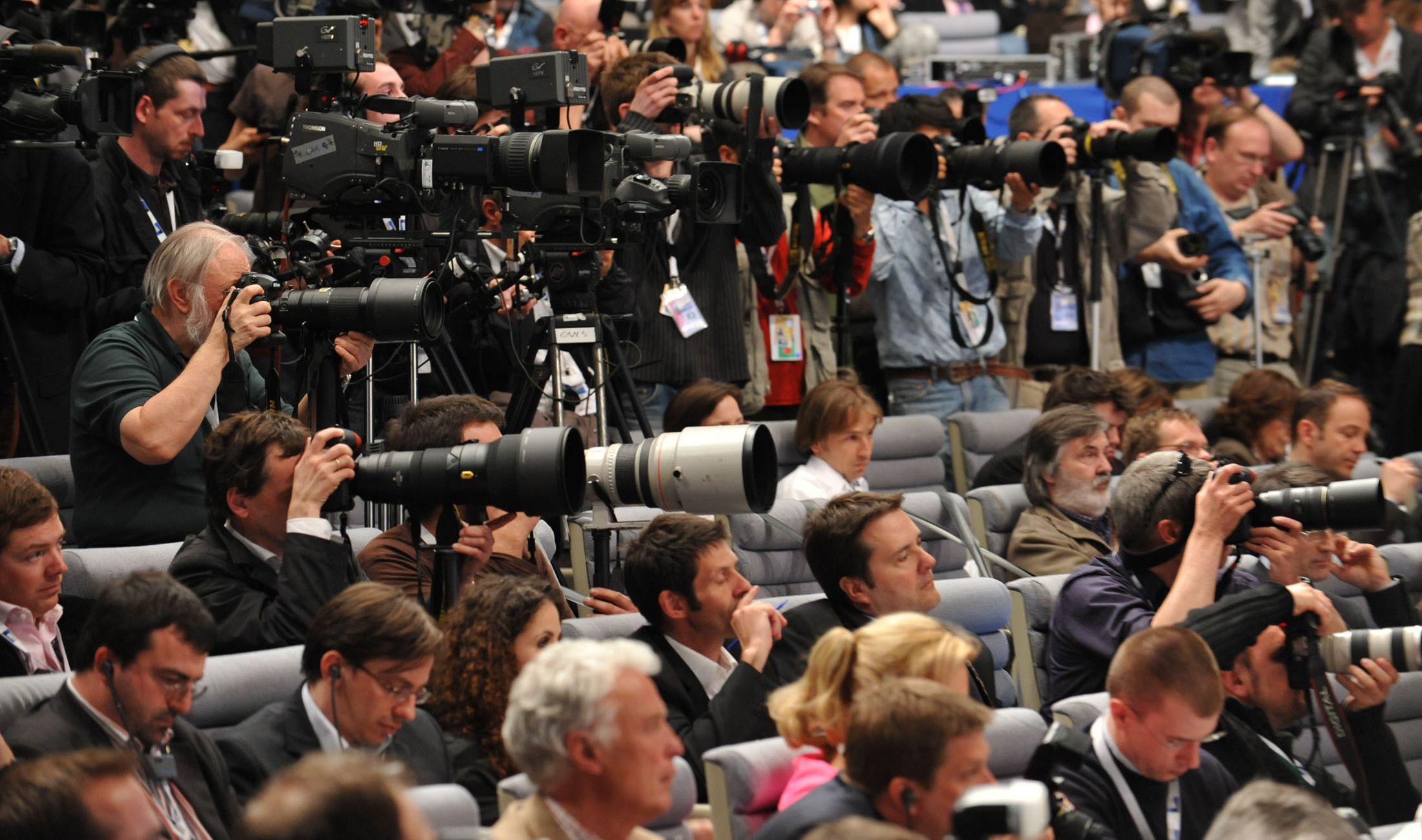 A large crowd of journalists and photographers capturing footage with cameras and lenses of varying sizes at an indoor press conference. The front rows are filled with people sitting and standing as they focus on the event.