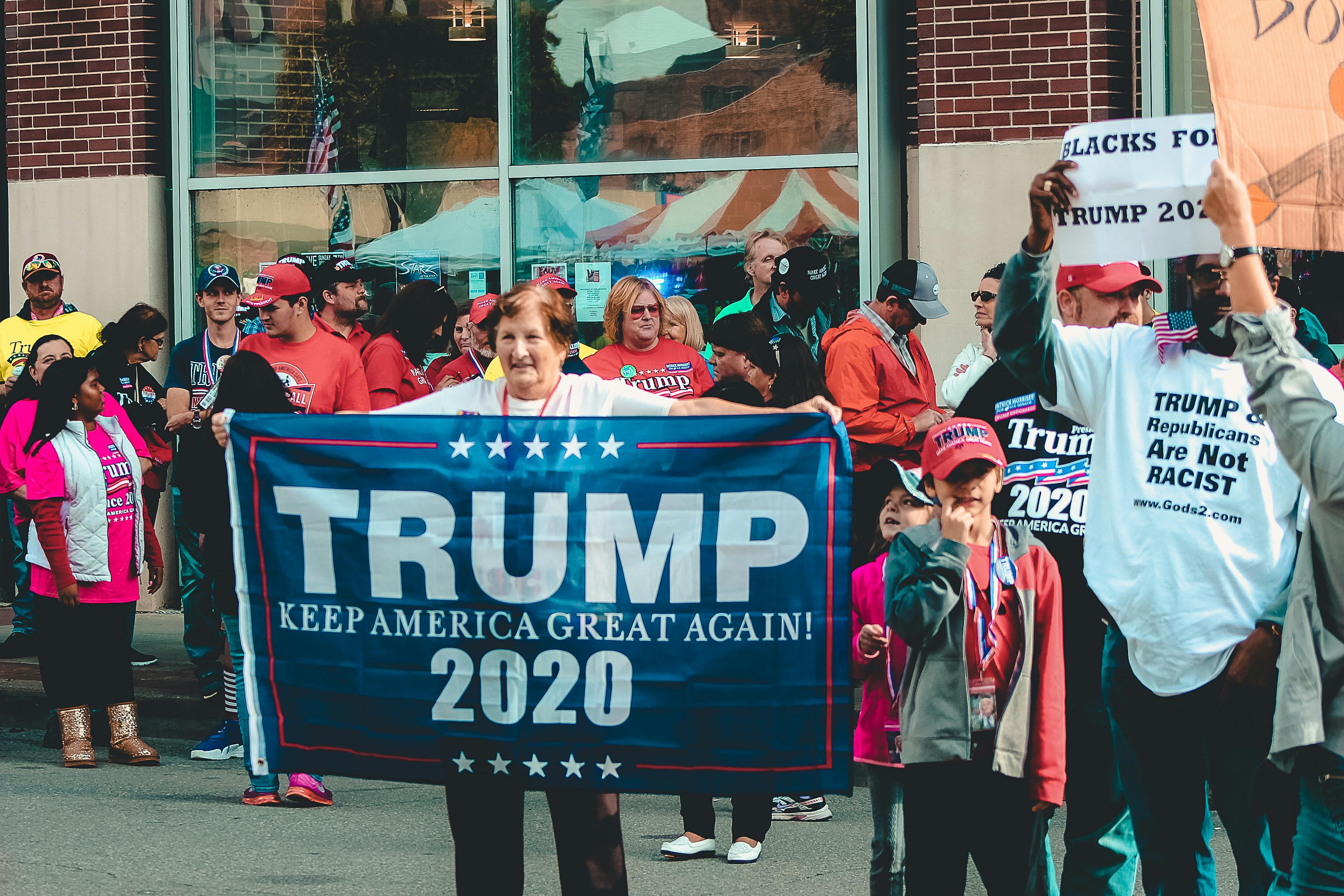 A group of people at a rally hold signs and banners supporting Trump 2020. Some wear red hats and shirts with political messages. They stand on a street in front of a building with large windows.