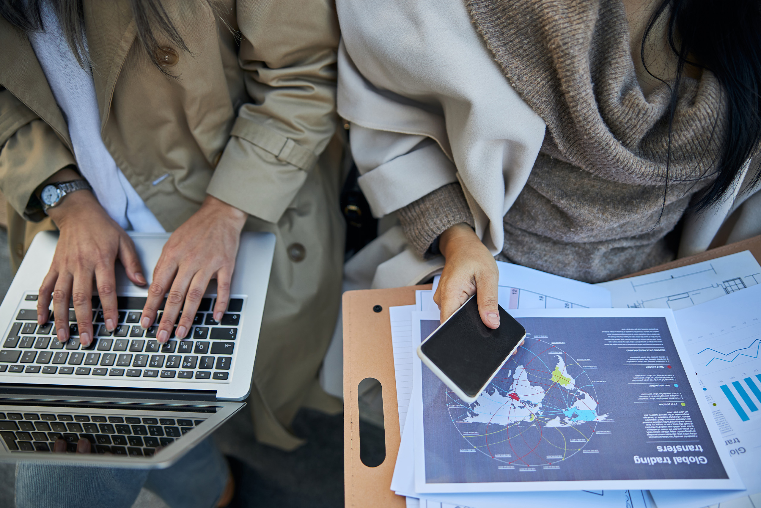 Two people sitting, one using a laptop, the other holding a smartphone. Documents with global trading graphs and charts are spread out on a clipboard. Both individuals are dressed in neutral-colored coats.