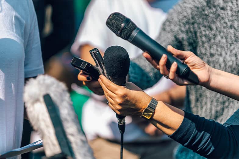 Several people hold various microphones and recording devices toward an unseen person, suggesting an interview or press conference setting. The focus is on the hands and equipment.