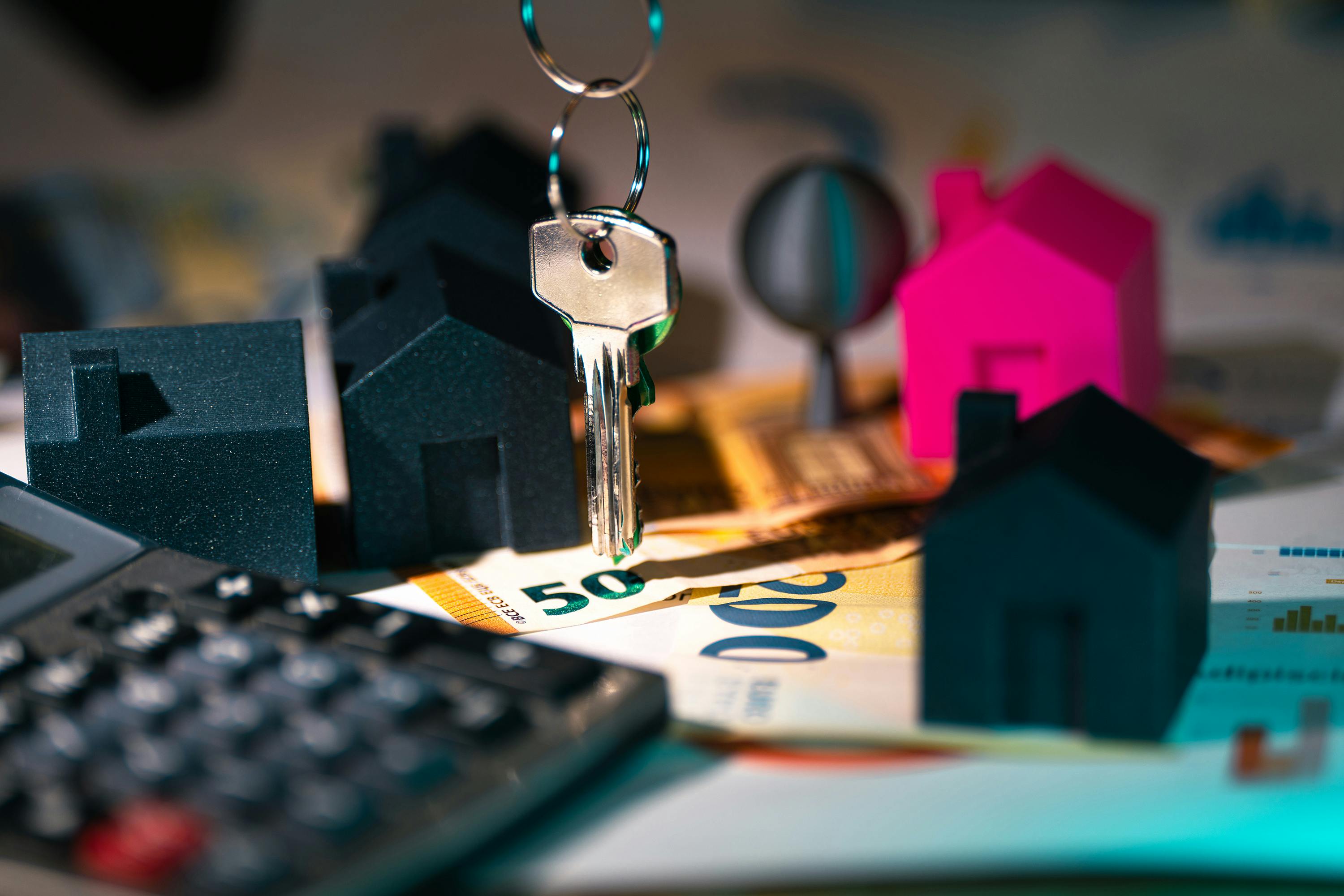 A close-up of miniature house models, a large key, and Euro banknotes on a table. A calculator is in the foreground, suggesting a financial or real estate theme with elements of home buying or investment.