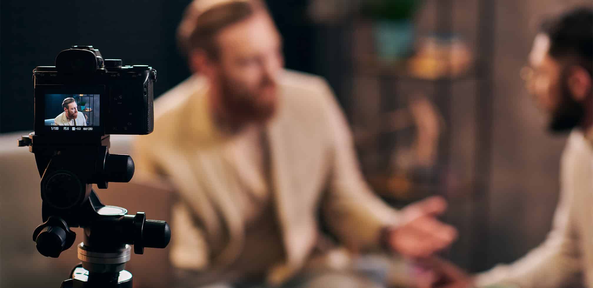 A camera on a tripod focuses on a blurry scene of two people seated and engaged in conversation. One person gestures with their hands. The room has a warm tone, and a shelf with plants can be seen in the background.